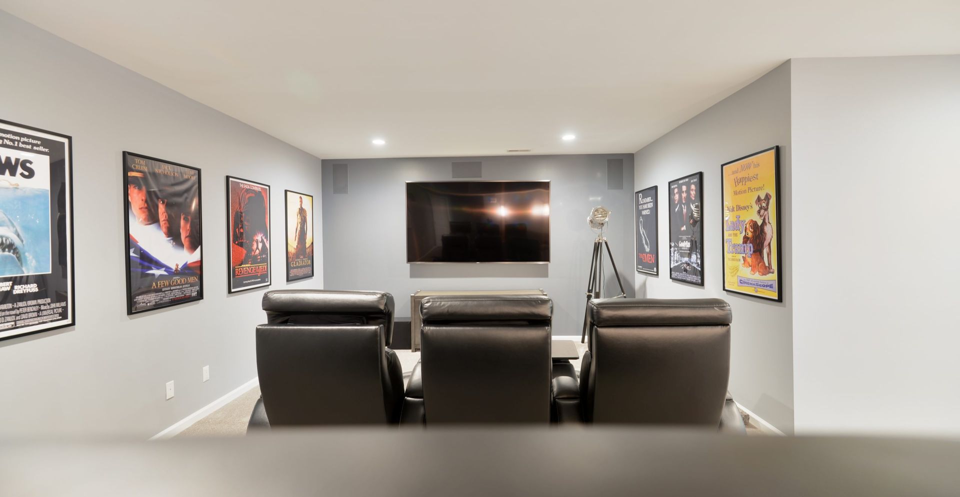 A home theater with three leather recliners facing a wall-mounted TV, surrounded by framed movie posters on gray walls.