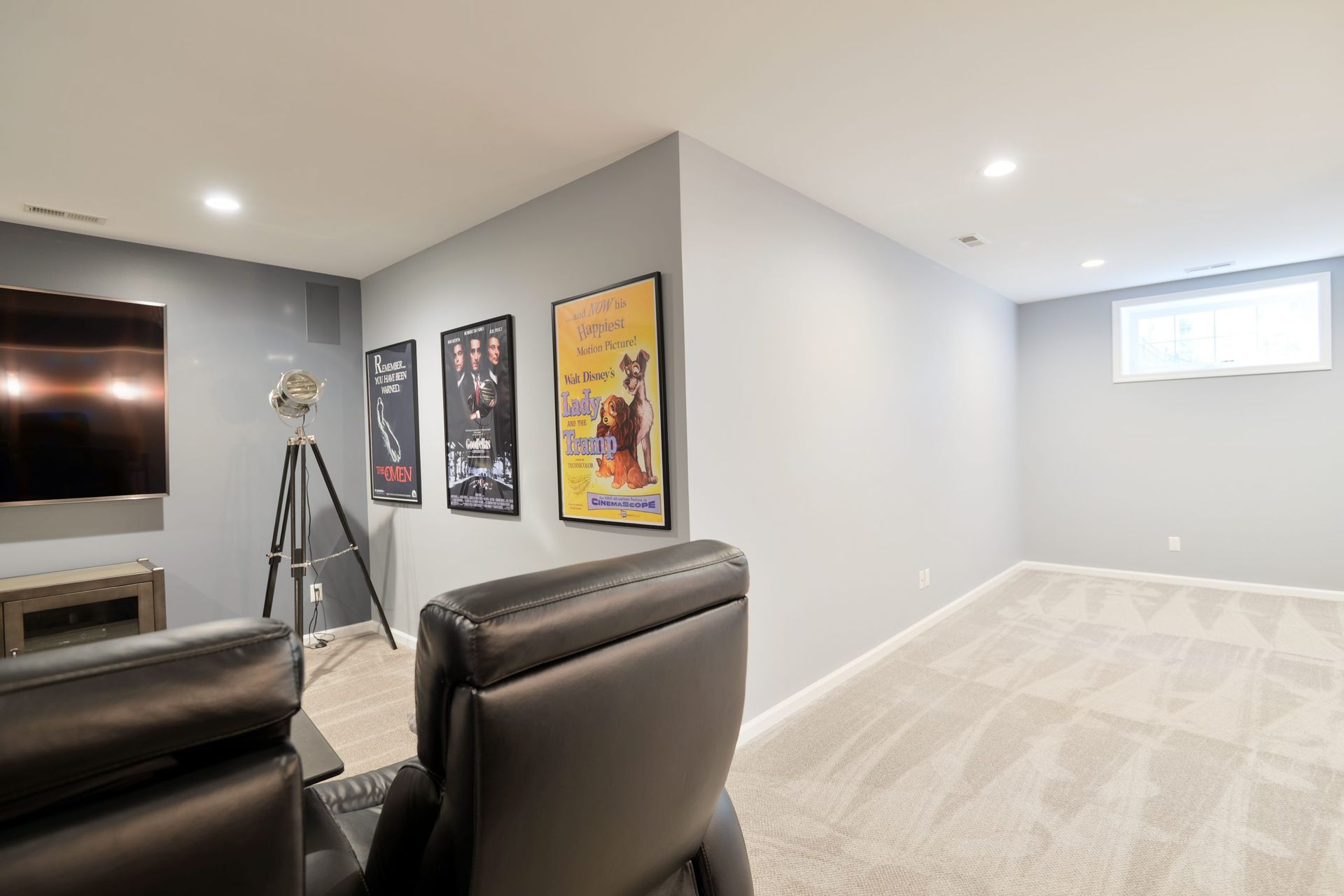 A basement media room featuring a large television, two black leather reclining chairs, and movie posters on the wall.