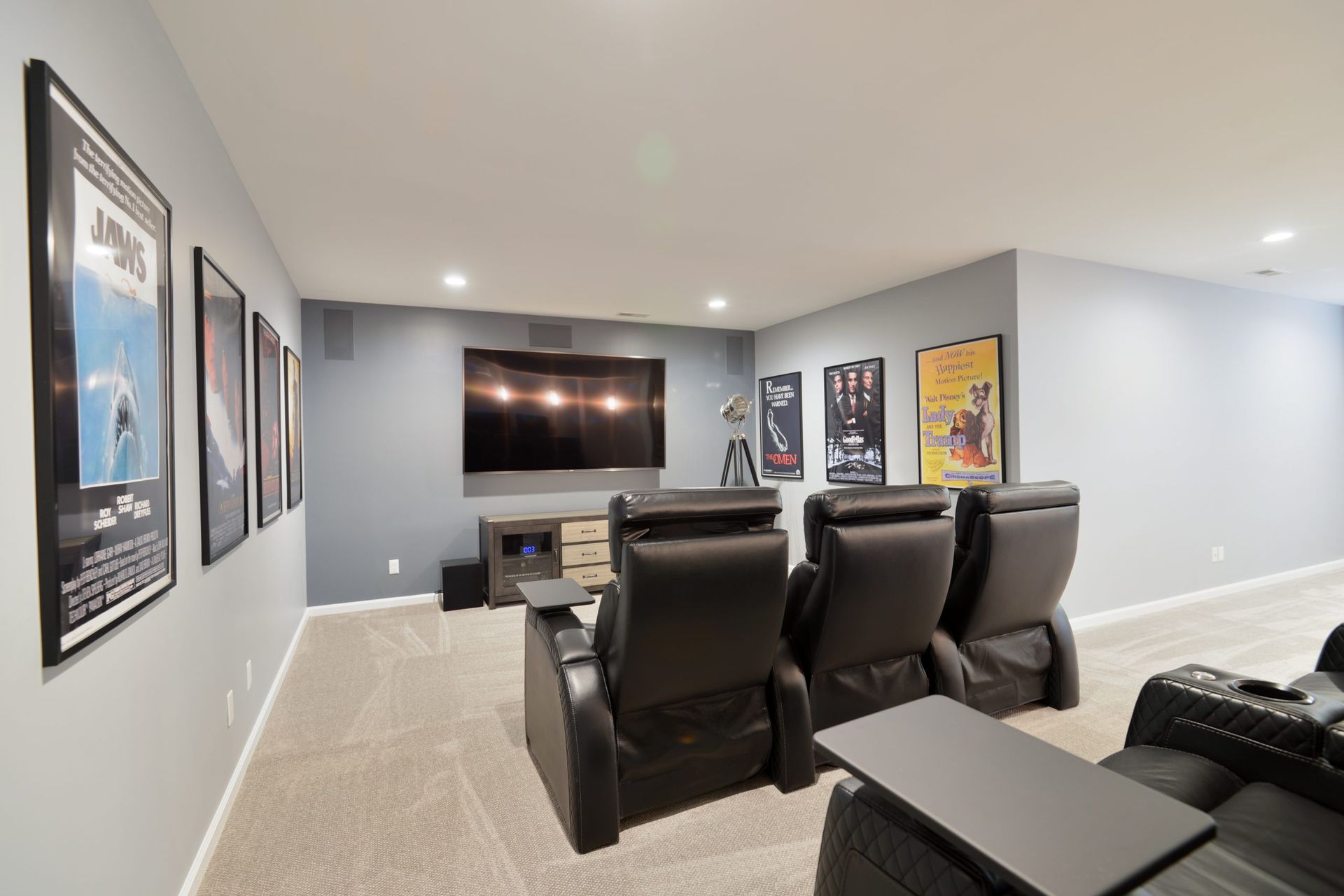 A home theater room with gray walls, carpeted floors, three rows of black leather recliners, and movie posters on walls.