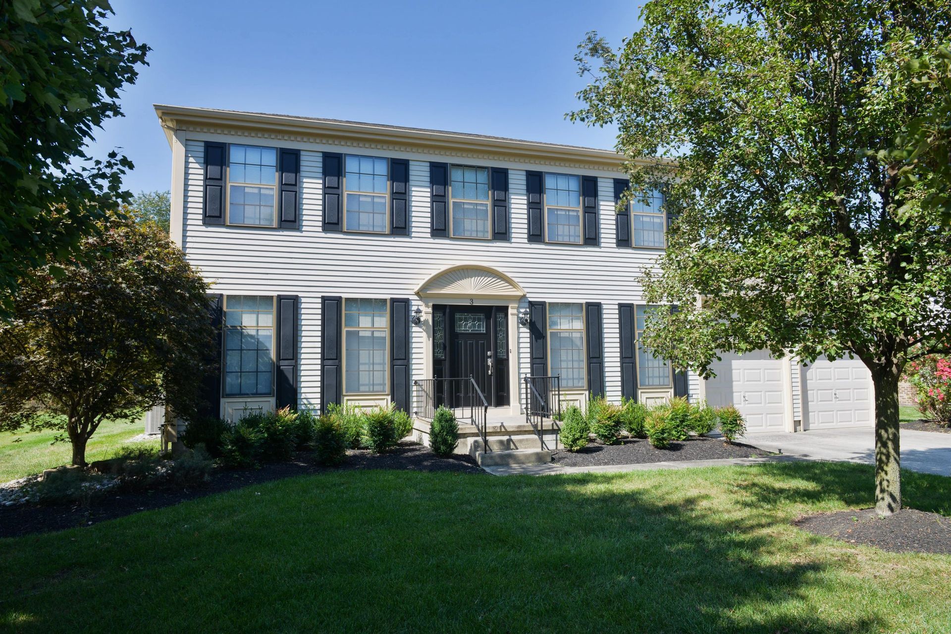 A two-story, light-colored suburban home with black shutters, a dark front door, and a front yard with trees and shrubs.