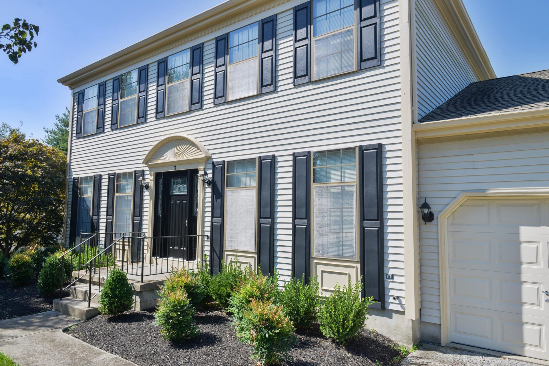 A two-story house with light-colored siding, dark shutters, and a front porch, viewed on a sunny day.