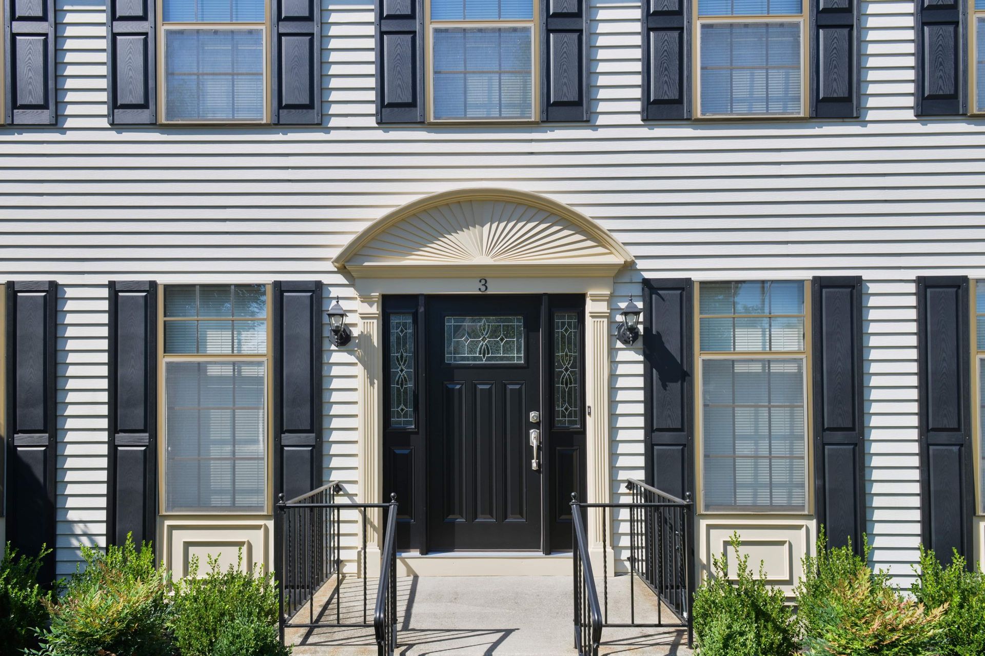 A house entrance with a black front door, arched decorative trim, surrounding windows with black shutters, and a walkway.