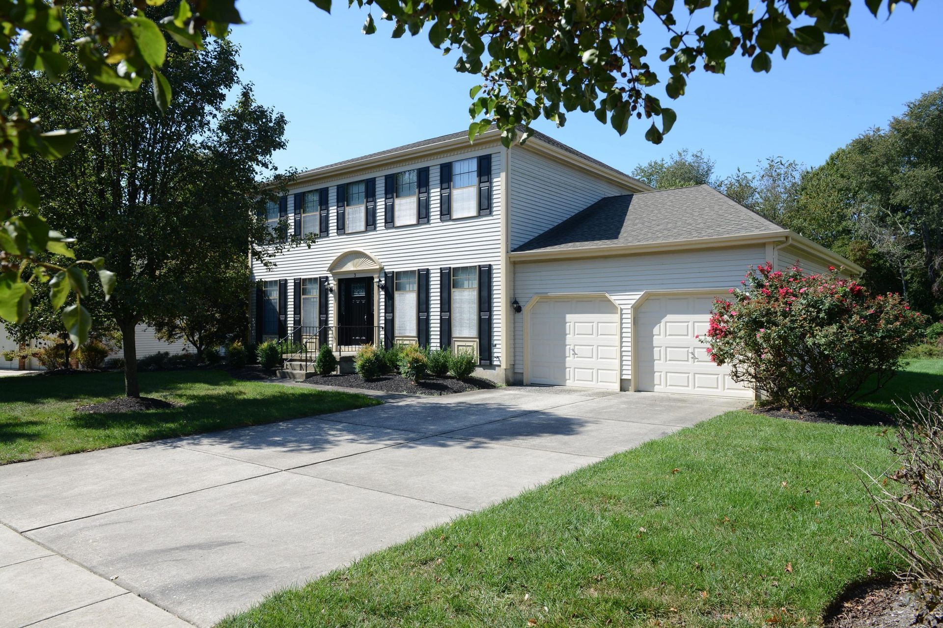 A two-story, light-colored suburban home with black shutters, a concrete driveway, and a two-car garage.