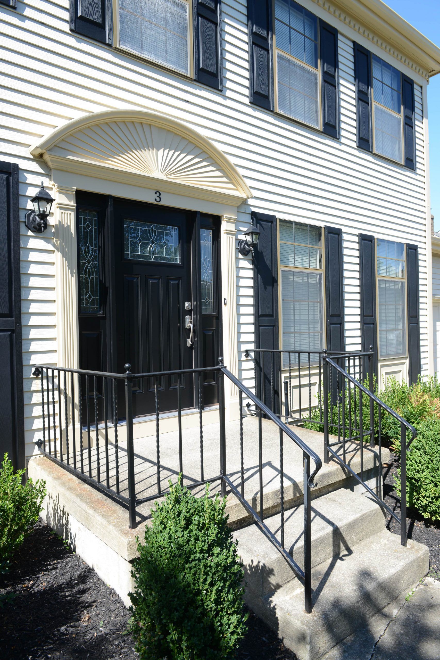 A front entrance of a two-story house with cream siding, black shutters, a dark front door, and a black metal railing.