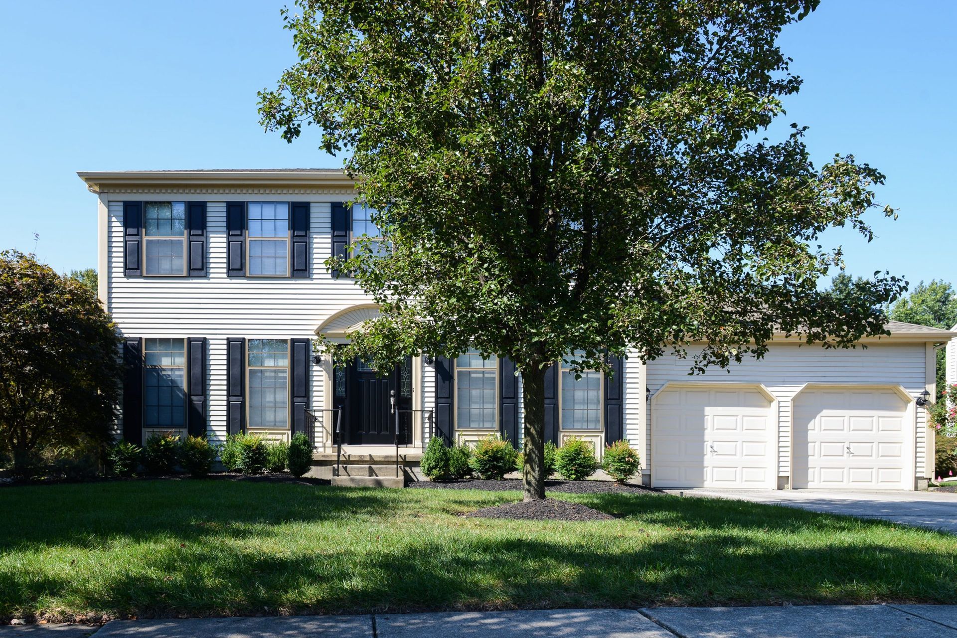 A two-story house with cream siding, dark shutters, a large front tree, and an attached two-car garage.