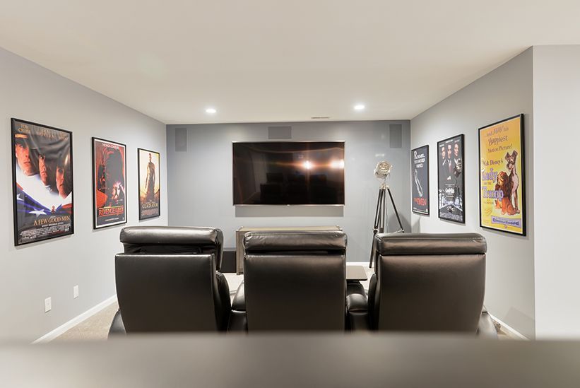 A home theater room with three dark leather recliner chairs facing a wall-mounted TV, decorated with framed movie posters.