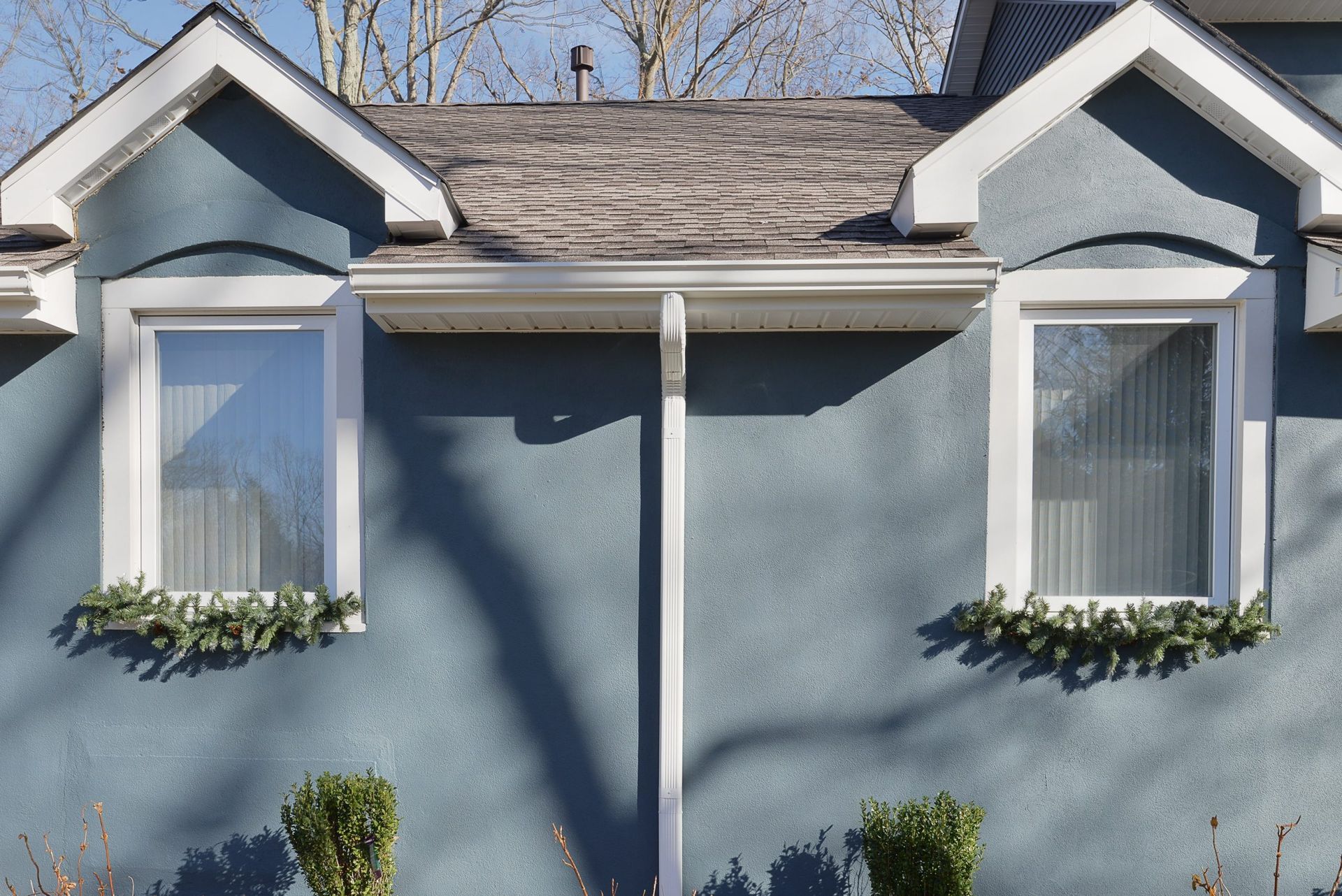 A blue, stucco-walled home exterior featuring two gabled windows with white trim, green window boxes, and a central downspout.