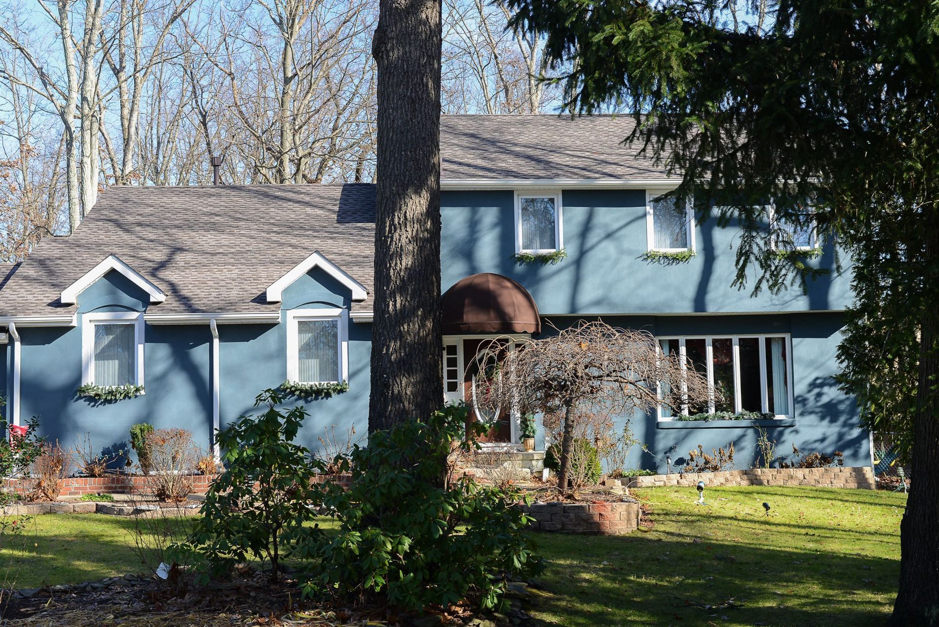 A two-story blue house with a gray shingled roof, brown arched entryway, and small landscaping in a wooded yard.
