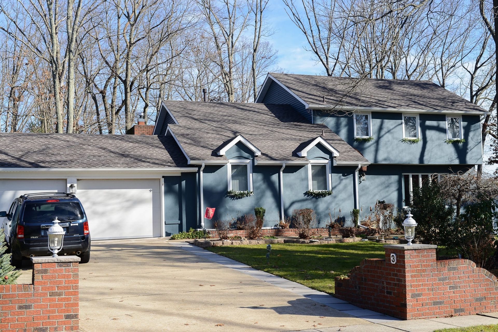 A blue two-story house with a white garage, gray shingled roof, and brick pillars at the edge of a paved driveway.