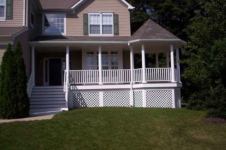 Tan house with a white front porch, stairs, railings, lattice skirting, and a small turret roof, surrounded by lawn.