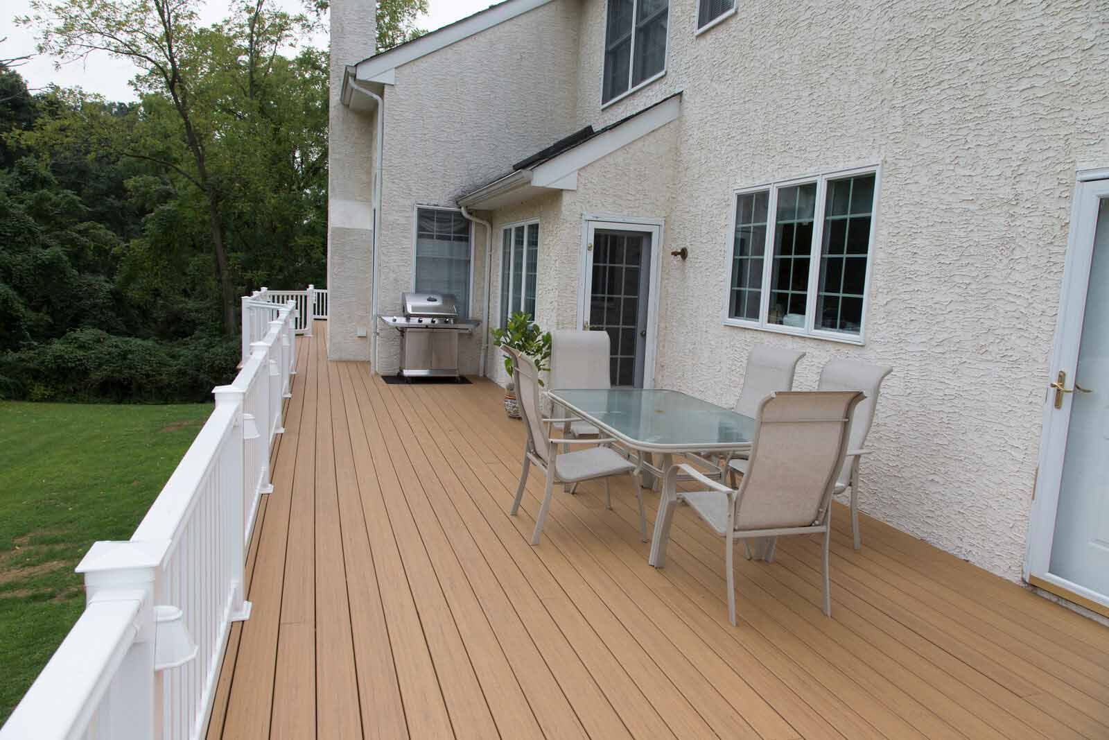 A tan composite deck with white railings, a patio dining set, and a grill, attached to a stucco-walled house.