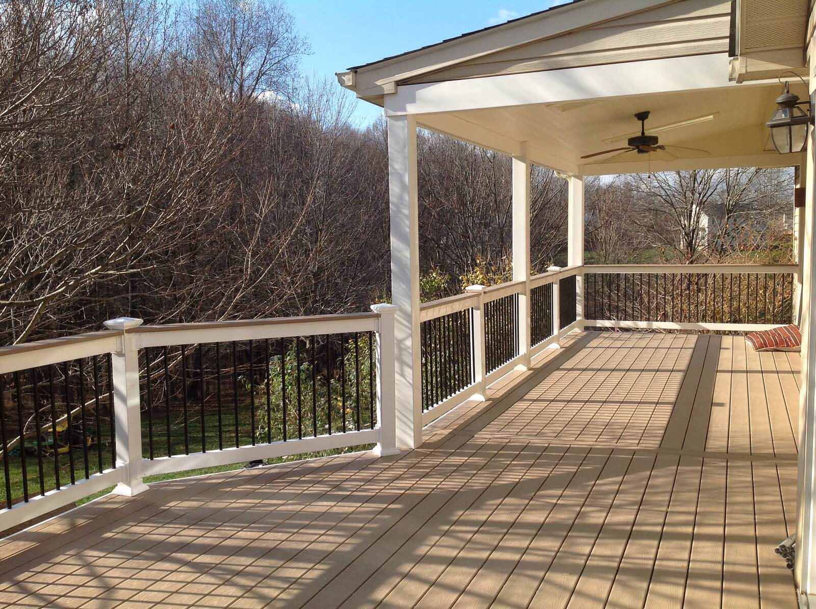 A covered wooden deck with a white railing and black spindles, overlooking a line of trees on a sunny day.