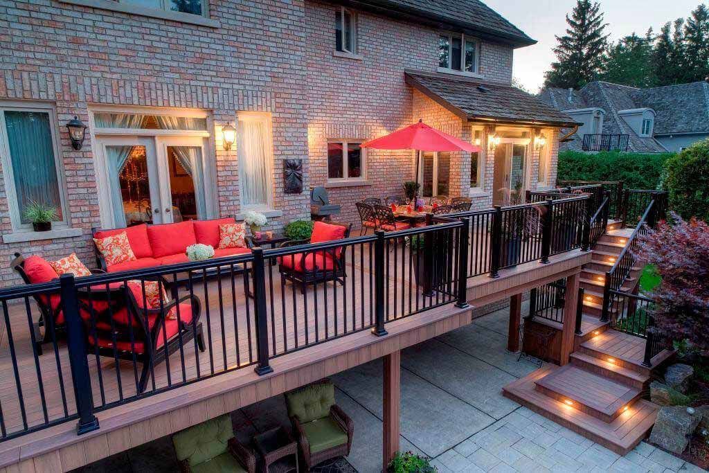 A multi-level wood deck with red outdoor furniture, a red umbrella, and lit stairs leading to a brick house at dusk.
