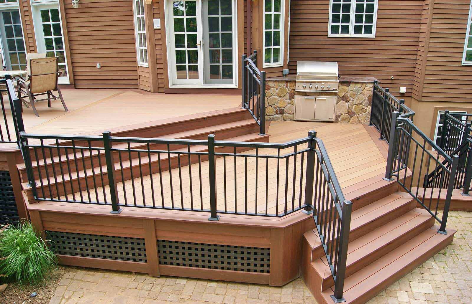 A multi-level wood deck with black metal railings, stone-encased grill area, and stairs leading down to a paver patio.
