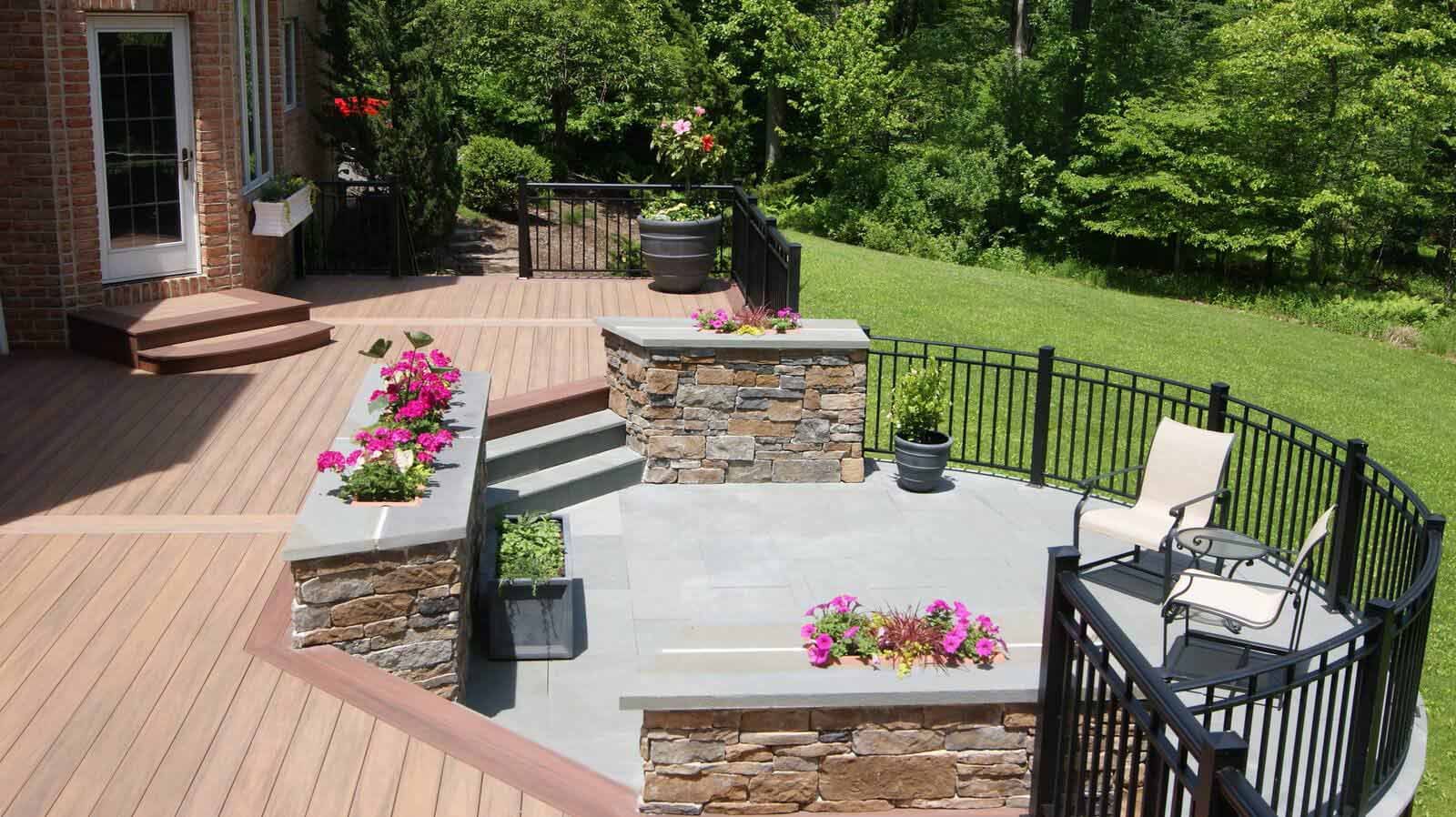 A patio with a light gray stone floor, stone retaining walls topped with flowers, and black railings overlooking a lawn.