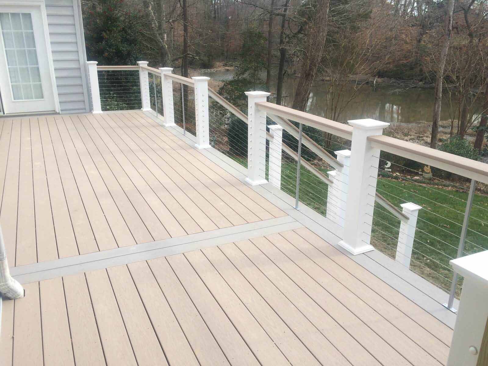 A beige composite deck with white railings and cable infill overlooking a wooded area and a pond.