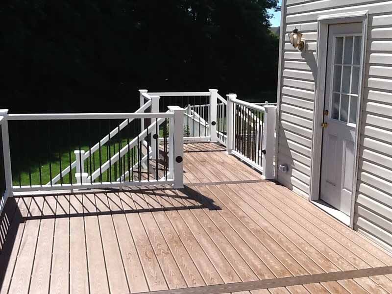 A wooden deck with white railings and black balusters leads to a doorway on a light-colored house.
