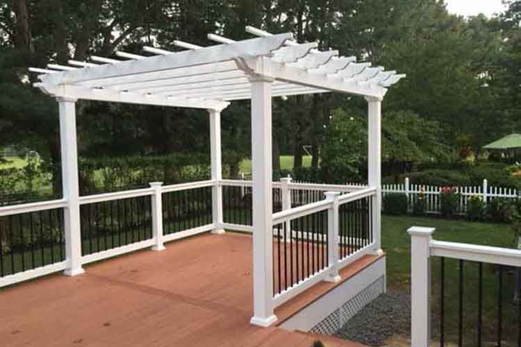 A white vinyl pergola stands on a raised wooden deck with white railings and black balusters in a grassy backyard.