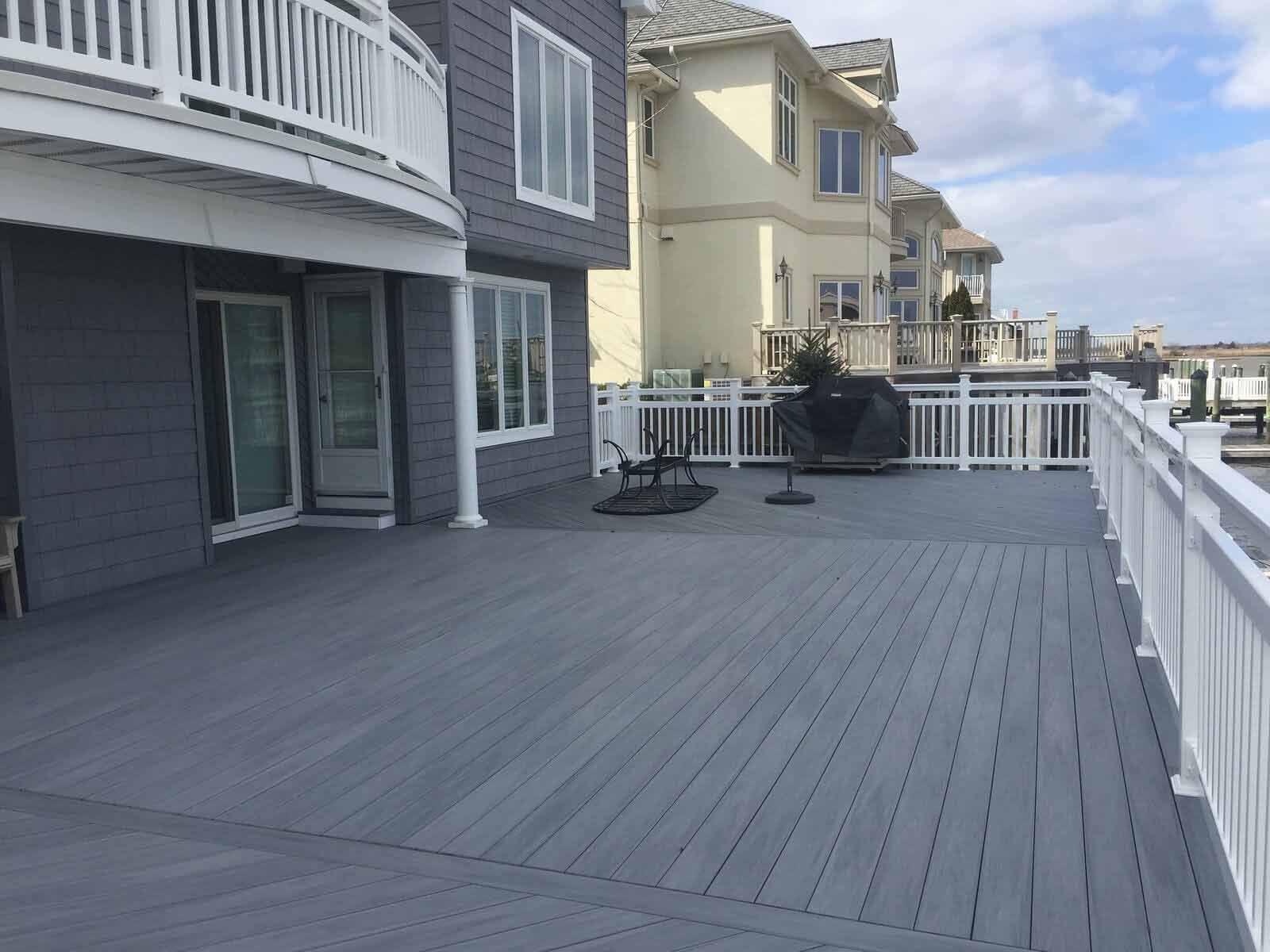 A large, gray composite deck with white railings attached to a two-story gray house next to a tan house by the water.