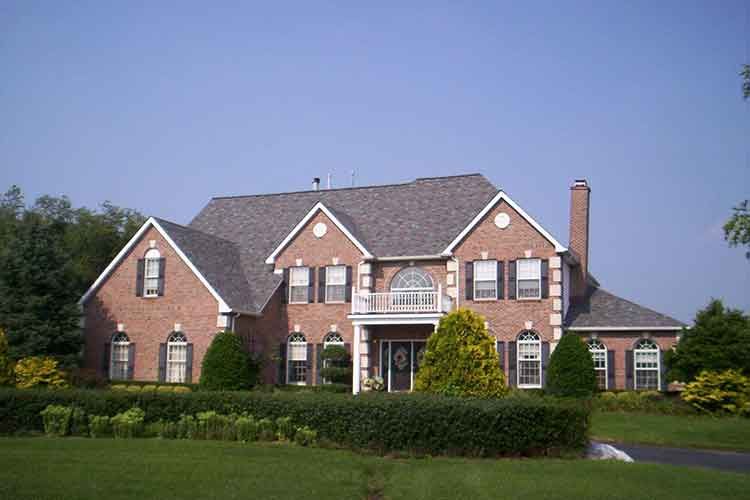 A large two-story brick house with a gray shingled roof, white trim, and a manicured hedge in the foreground.