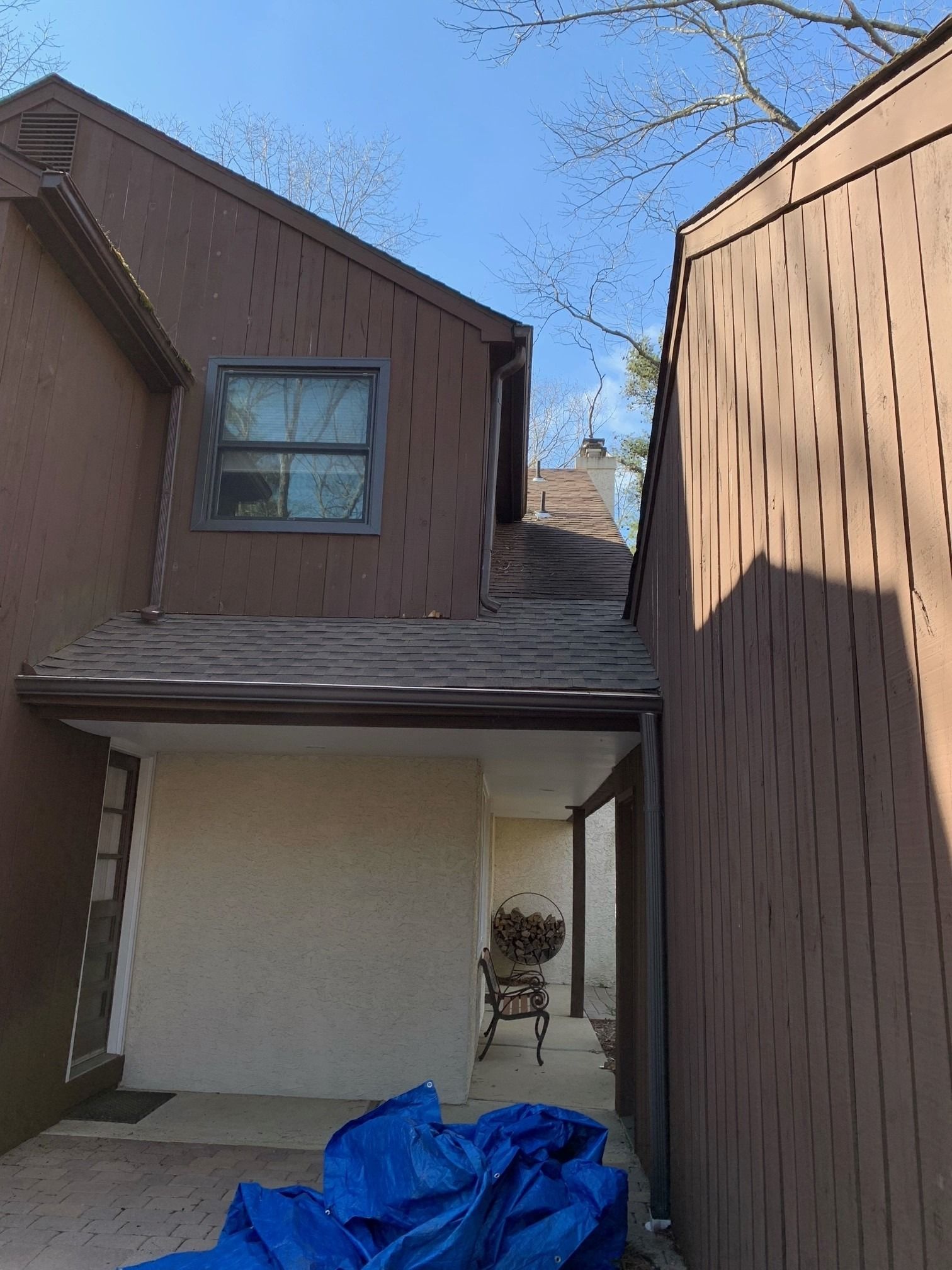 A view of a brown wooden-sided house exterior with a small patio, a window on the upper level, and a blue tarp in front.