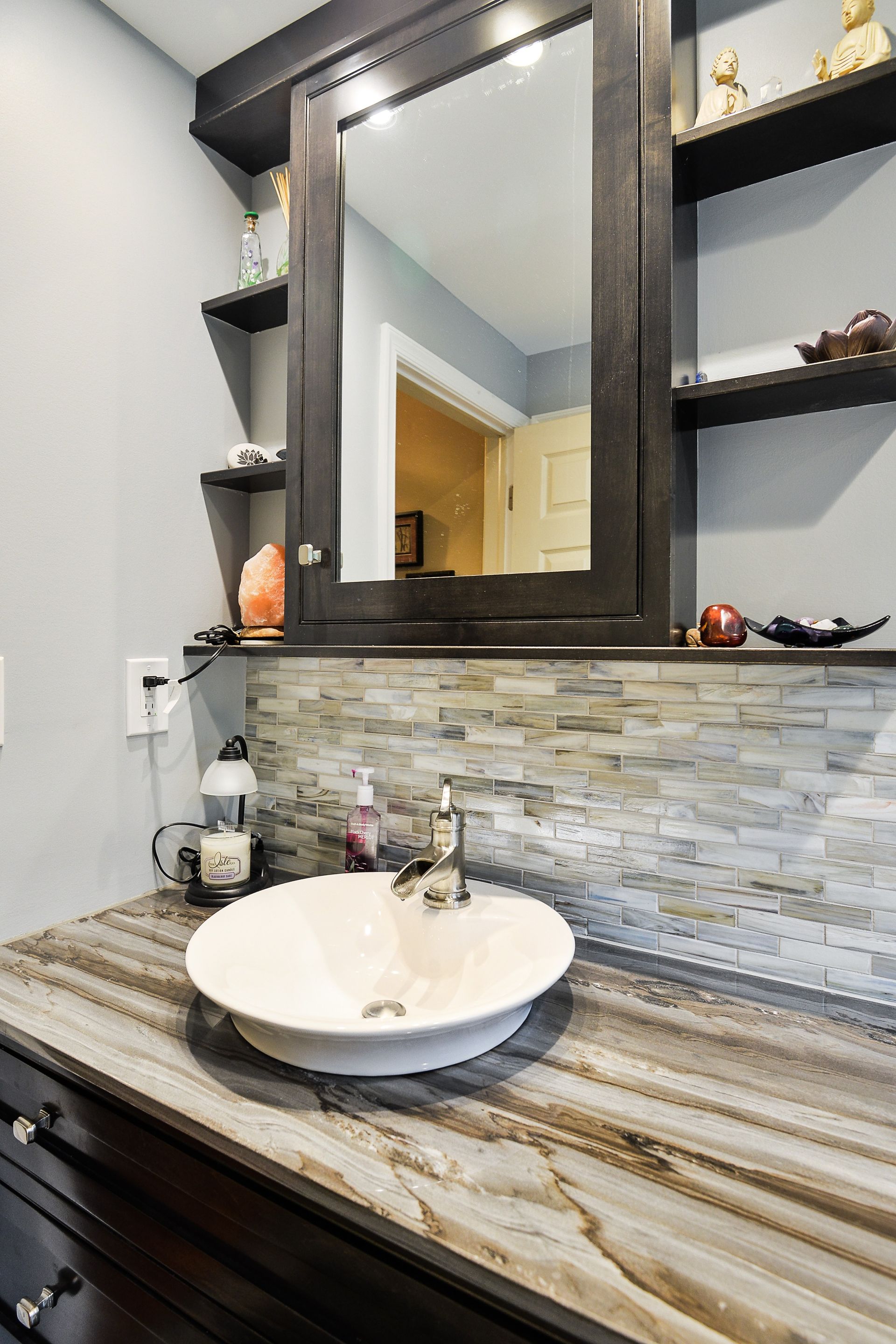 A modern bathroom vanity with a white vessel sink, marble-patterned countertop, stone tile backsplash, and dark mirror.