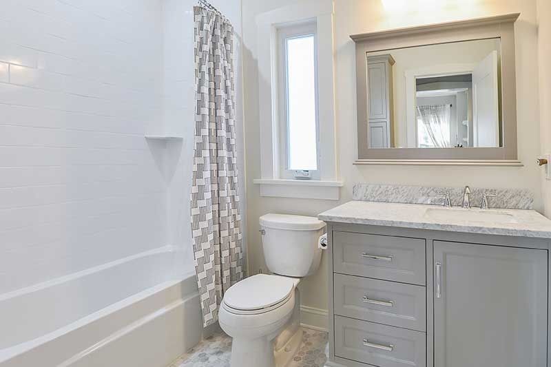 A bright bathroom with a white bathtub, toilet, gray vanity with a mirror, and a gray-and-white patterned shower curtain.