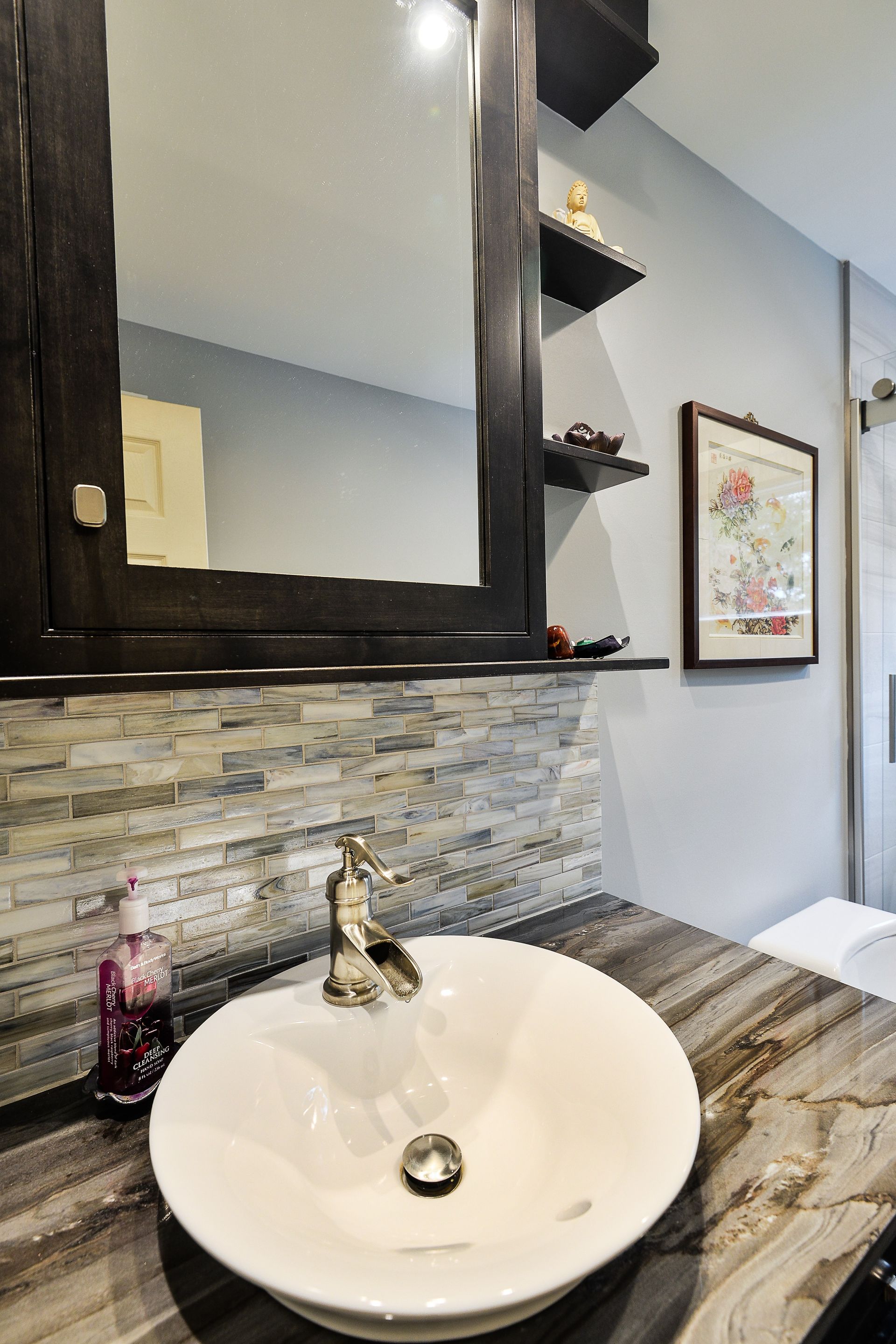 A bathroom vanity with a white vessel sink, ornate gold faucet, stone tile backsplash, and a dark wood mirror.