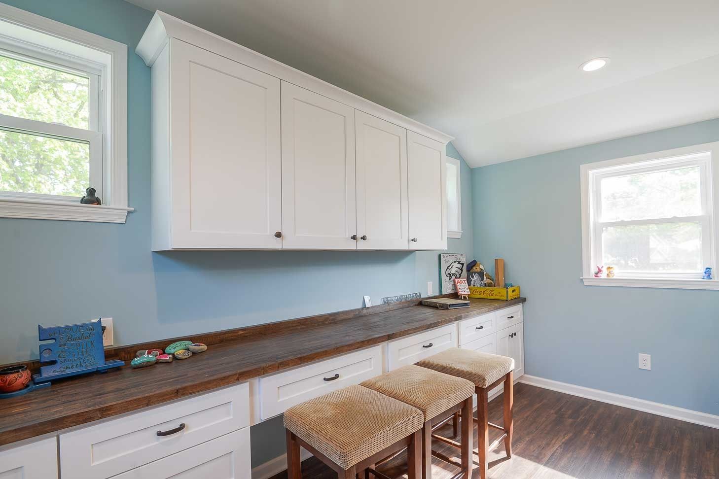 A desk area with white cabinetry, a dark wood countertop, and three woven stools against a light blue wall.