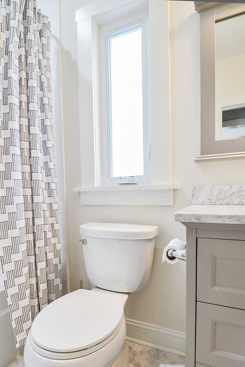 A modern bathroom featuring a white toilet, gray vanity with a marble top, and a patterned shower curtain beside a window.