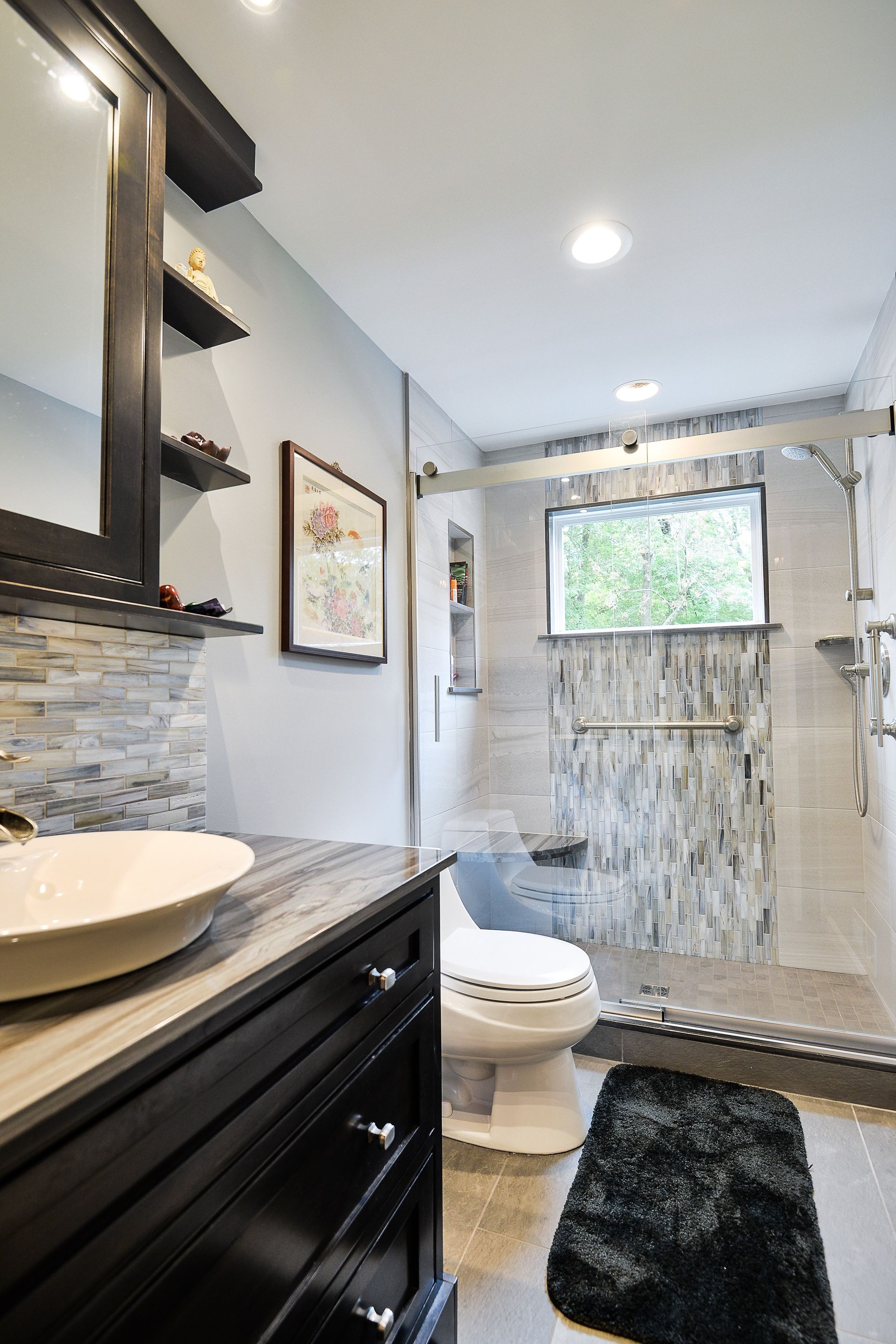 A modern bathroom with a dark vanity, vessel sink, framed art, a toilet, and a glass-enclosed shower with tiled walls.