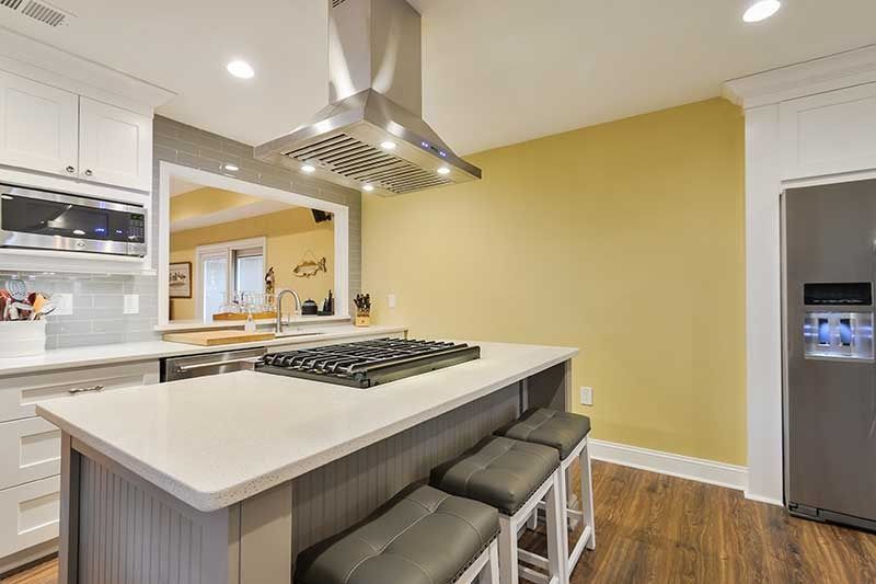 A kitchen with a center island featuring a gas stovetop and three grey stools, next to a stainless steel refrigerator.