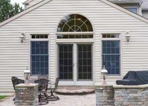 A beige vinyl-sided house exterior with a patio, arched transom window over French doors, and stone pillars with lanterns.