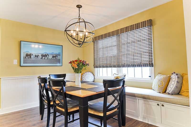 A yellow dining nook with a dark wood table, black chairs, a geometric chandelier, and built-in bench seating by a window.