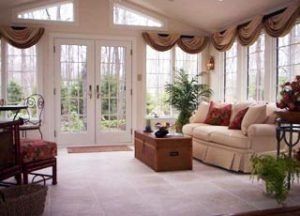 A sunroom with a beige sofa, a wooden trunk coffee table, large glass doors, and draped curtains overlooking a forest.