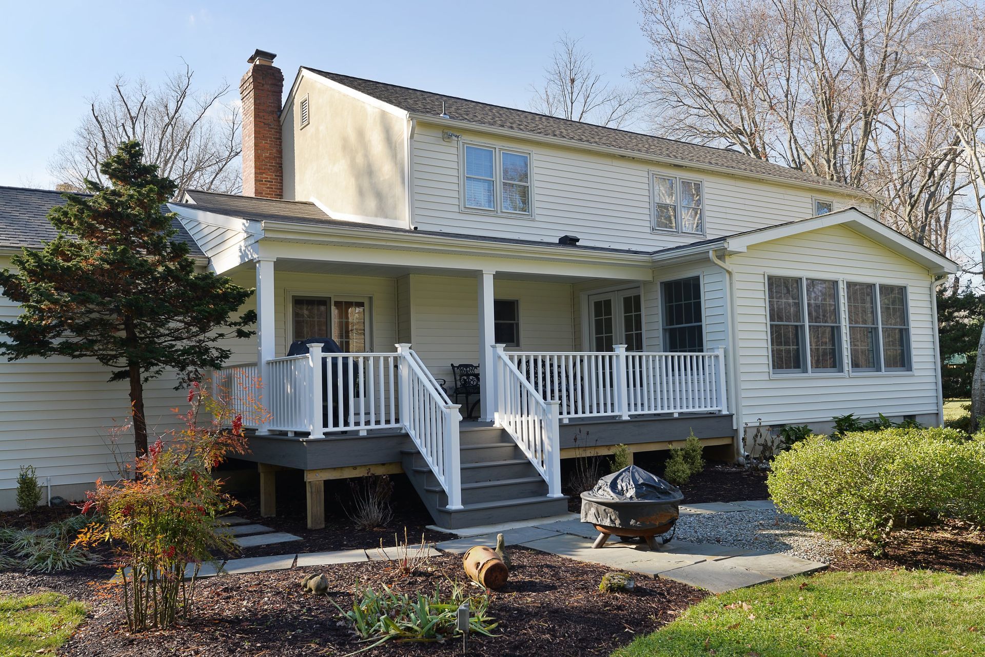 A two-story white house with a gray deck, white railing, stairs, a fire pit, and landscaping on a sunny day.