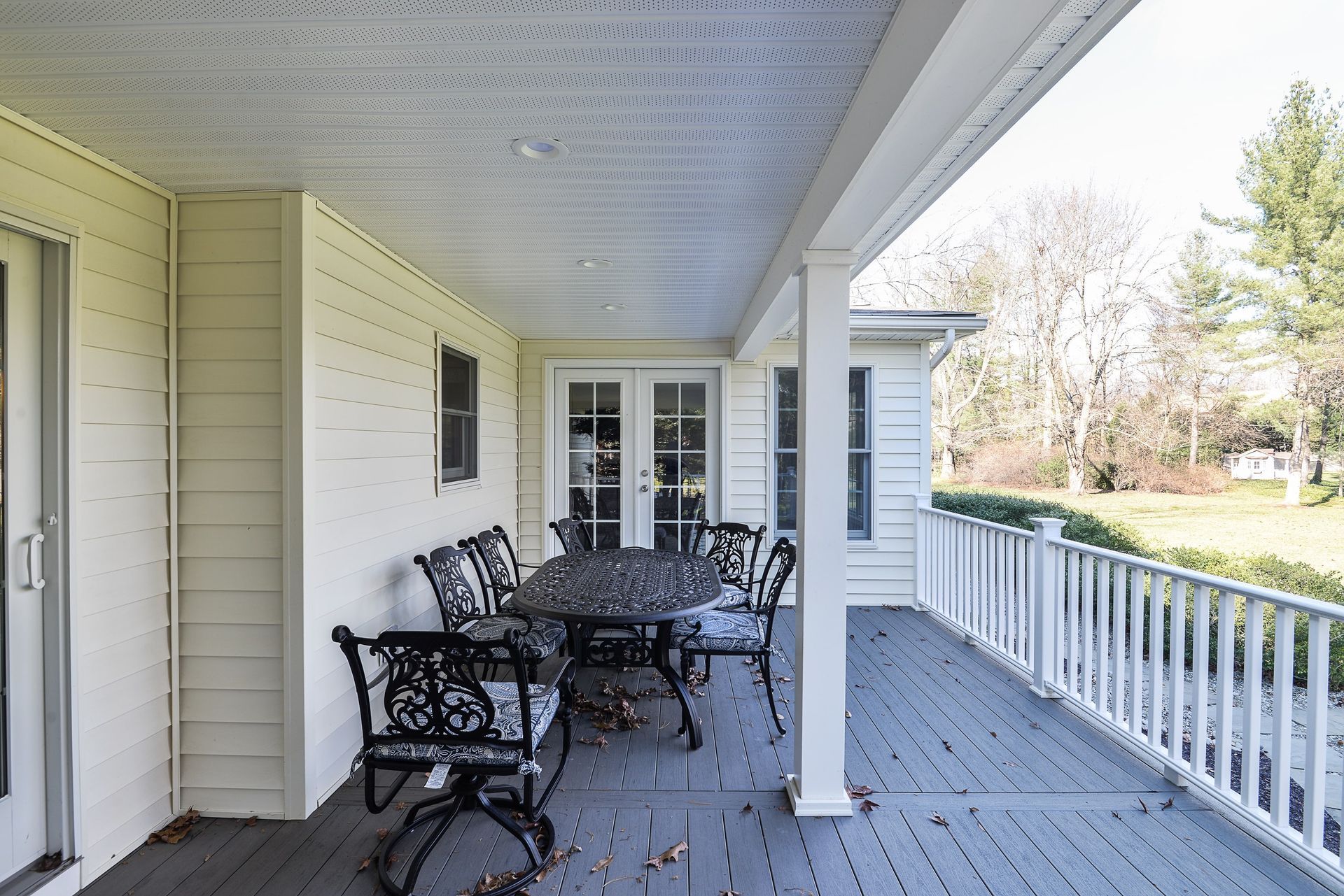 A covered outdoor porch with a dark metal dining set on grey decking, facing a grassy yard with trees.
