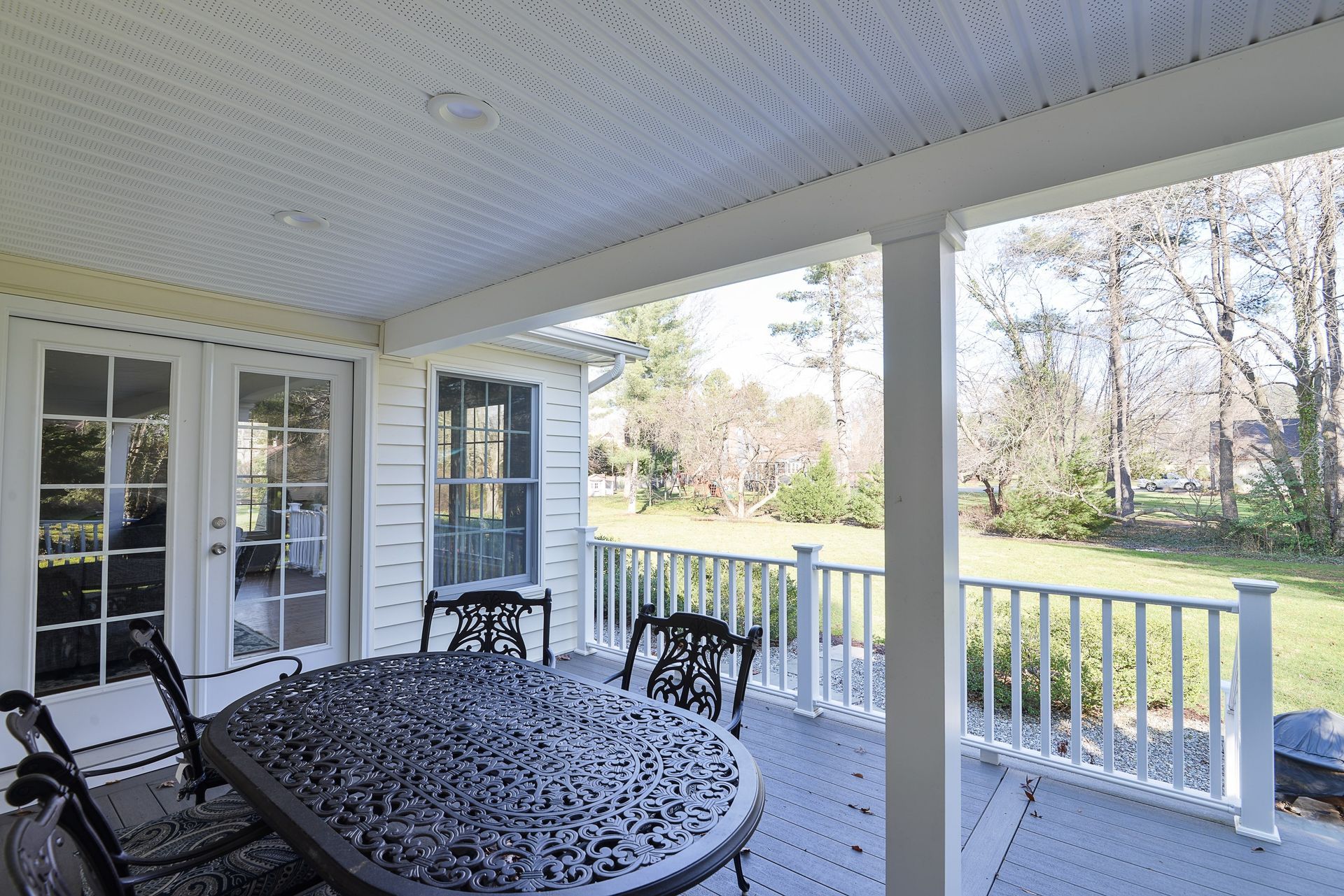 Covered outdoor deck with white railing, a black metal table and chairs, looking out onto a backyard with trees.