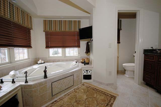 A bathroom with a jetted corner tub, wood blinds, tile flooring, a rug, and a separate doorway leading to a toilet.