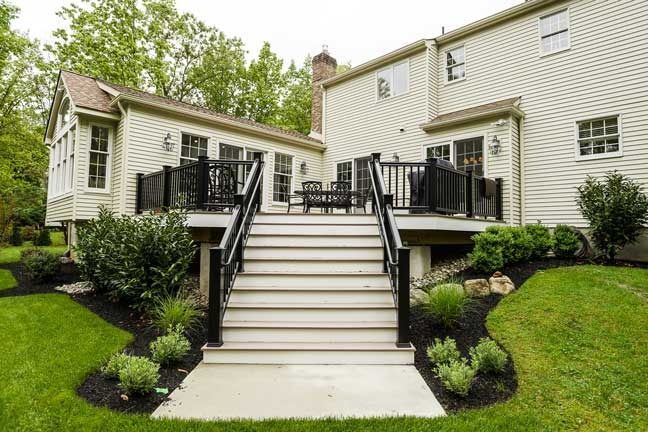 A wide, central staircase leads up to a raised outdoor deck on the back of a light-colored, two-story house with a lawn.
