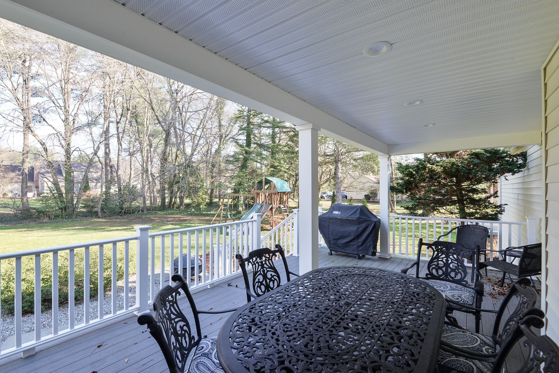 A patio with a dark metal dining set and grill, overlooking a green backyard with trees and a playset.