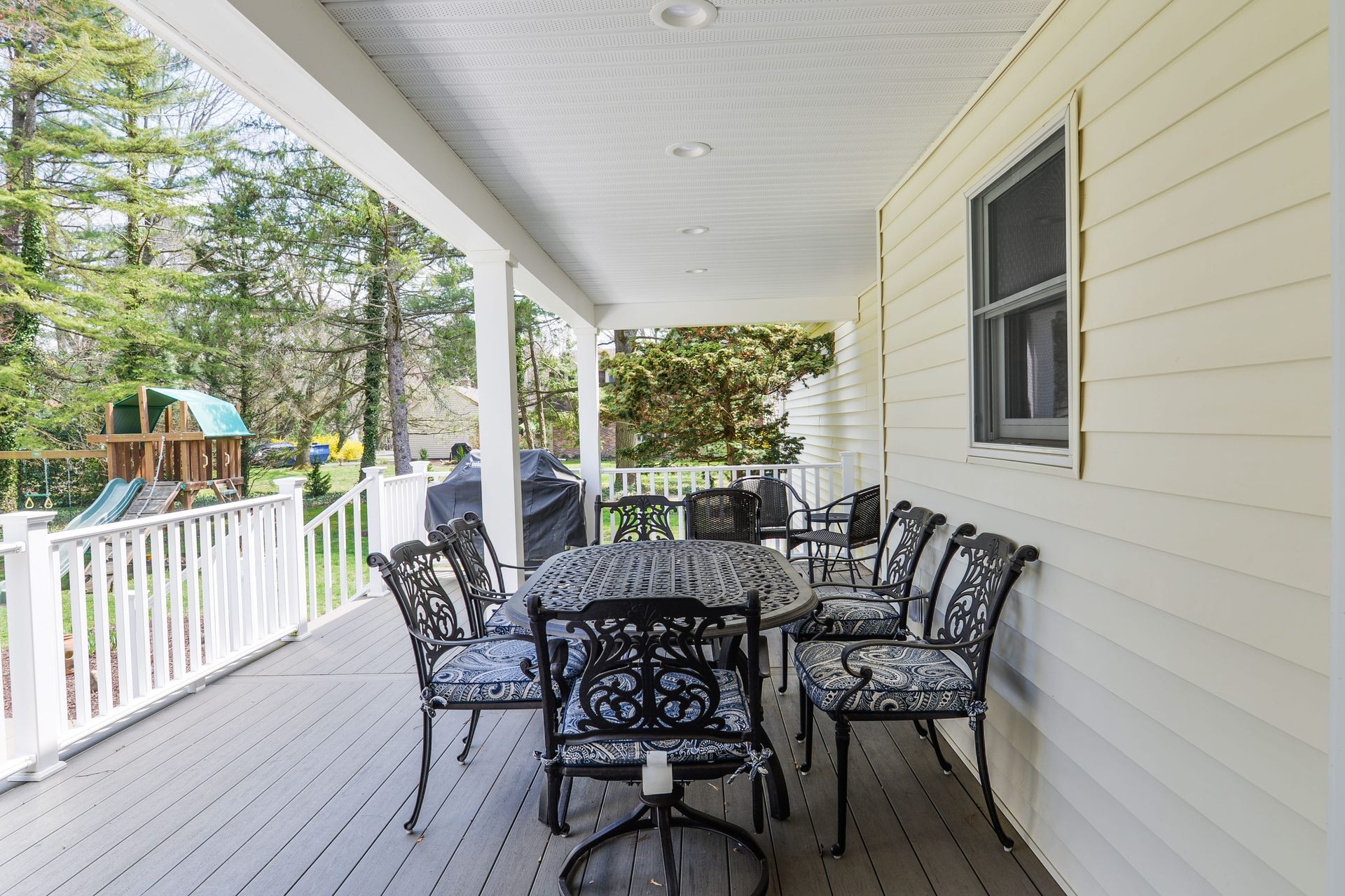 A covered outdoor porch with light yellow siding, featuring a metal dining set and a view of a backyard with a playset.