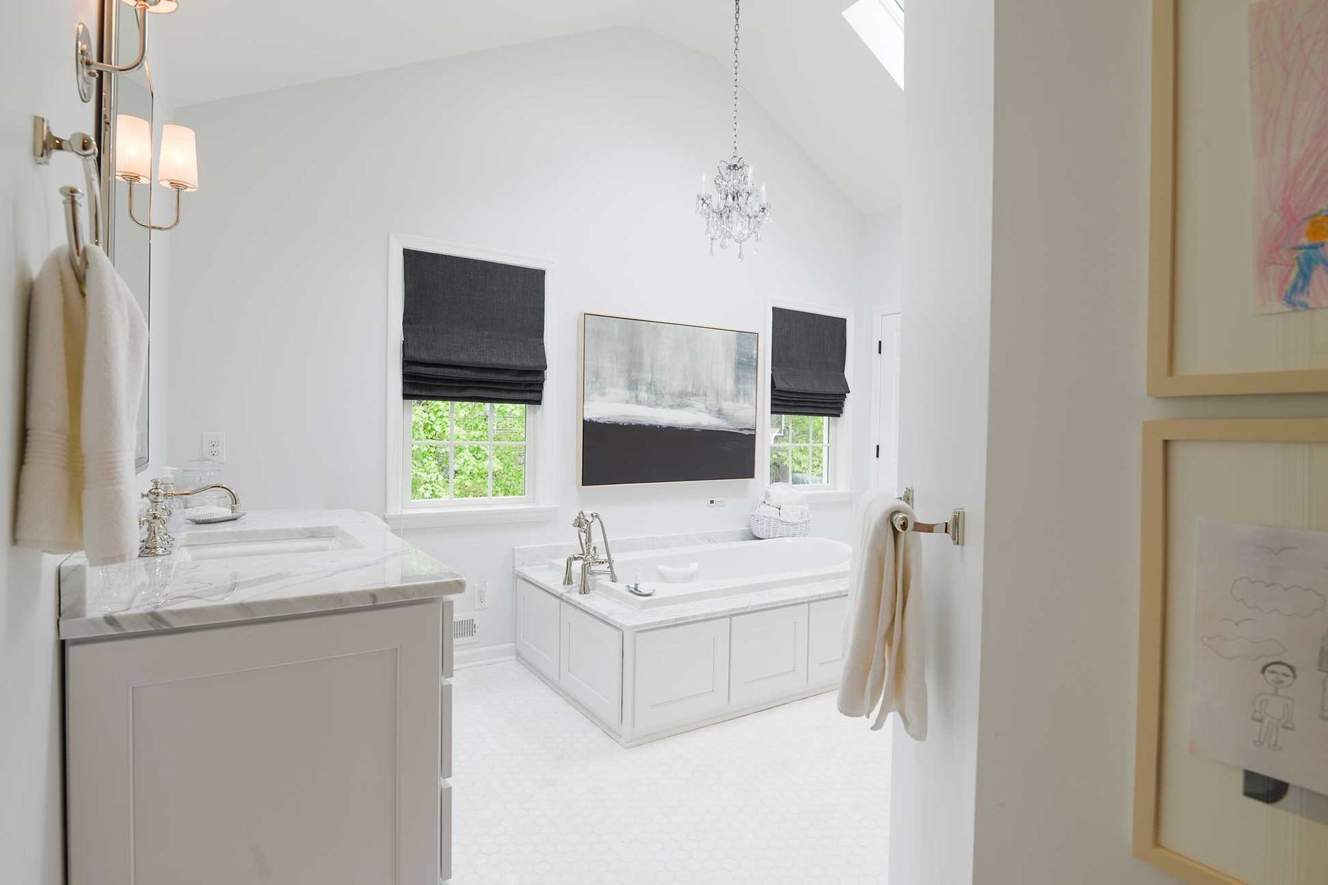 A white bathroom with a deep soaking tub, a marble vanity, black roman shades, and a chandelier under a vaulted ceiling.