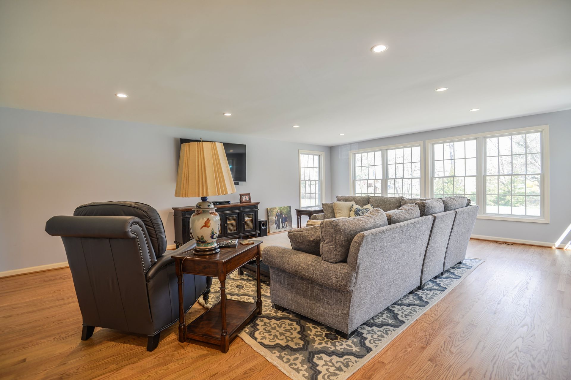 A living room with light blue walls, hardwood floors, a gray sectional sofa on a patterned rug, and a dark armchair.
