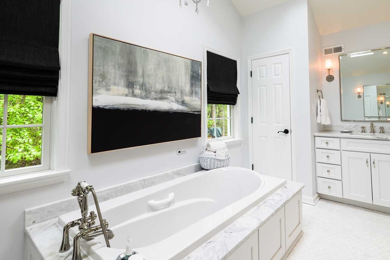 A bright, modern bathroom featuring a white soaking tub with marble trim, black shades, and a large abstract painting.