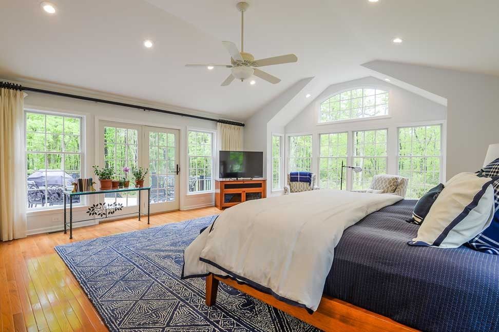 Spacious bedroom with a large bed, navy rug, wooden flooring, and a ceiling fan, featuring multiple windows viewing trees.