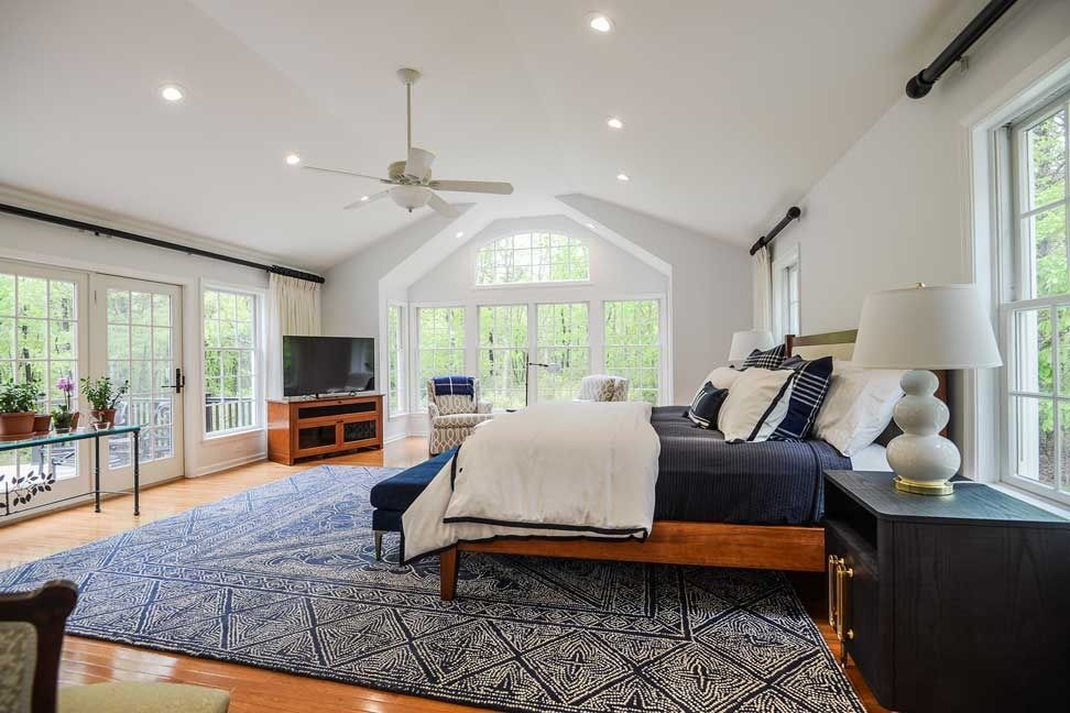 A spacious bedroom with a king bed, blue patterned rug, wooden furniture, ceiling fan, and large windows with garden views.