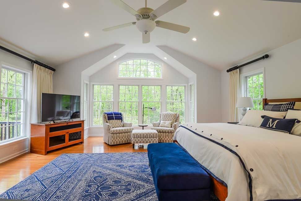 Bright bedroom with a large bed, navy bench, blue patterned rug, two armchairs, and windows overlooking woods.