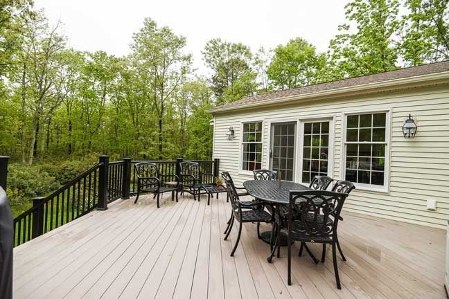 A light-colored wooden deck with a black patio dining set, featuring a railing overlooking a lush forest.