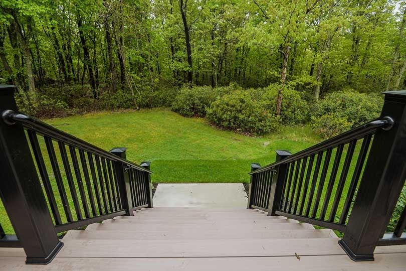 A view from a wooden deck looking down stairs toward a green grassy backyard bordered by a dense forest.